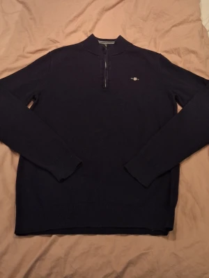 Mörkblå halvzip tröja från Gant - Mörkblå stickad tröja från Gant med halv dragkedja och liten broderad logga på bröstet. Tröjan har ribbade muddar vid ärmslut och nederkant samt en klassisk ståkrage. Perfekt för en stilren och avslappnad look. Passar dig runt 150-160cm!