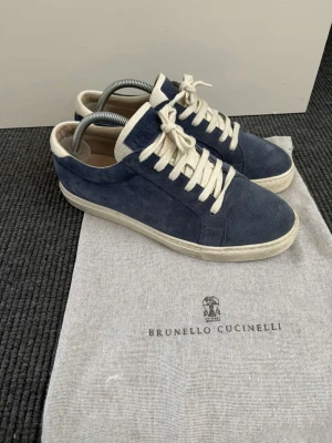 Brunello Cucinelli sneakers - Stilrena sneakers i blå mocka från Brunello Cucinelli med vita detaljer och snörning. Skorna har en klassisk låg profil och vit sula, perfekta för en clean och avslappnad look. Kommer med originalpåse.