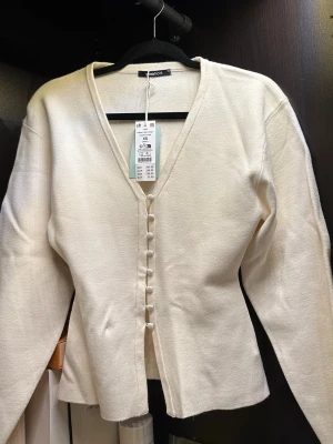 Gina Tricot långärmad kofta med knappar  - Helt ny oanvänd cardigan / långärmad blus med v-ringning och tygklädda knappar framtill. Koftan har lång ärm och lätt figursydd passform. Storlek XS vit aldrig använd prislapp kvar. 