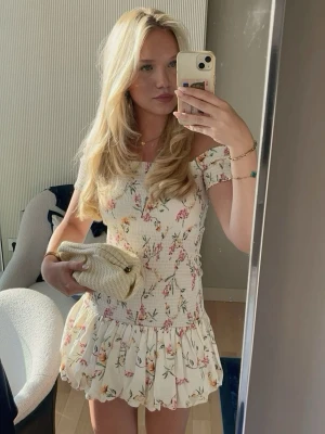 Blommig offshoulder kortklänning - Supersöt kortklänning med offshoulder och volangkjol. Klänningen är vit med blommigt mönster i rosa, grönt och gult. Smockad överdel och axelbandslös design ger en feminin och somrig känsla. Perfekt för varma dagar och picknickar.