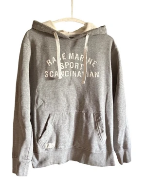 Grå hoodie från Race Marine - Grå hoodie från Race Marine med vit text på bröstet: 'RACE MARINE SPORT SCANDINAVIAN'. Klassisk känguruficka framtill, huva med vita dragsnören och ribbade muddar. Mjuk bomullsblandning som är skön att bära.