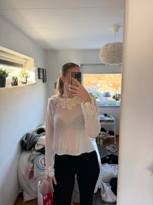 Vit lätt stickad tröja med blommor  - Söt vit blus i transparent material med långa ärmar. Blusen har dekorativa virkade blommor runt halsringningen och vid ärmsluten. Helt oanvänd 