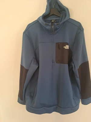 Blå hoodie från The North Face - Snygg blå hoodie från The North Face med dragkedja i halsen och bröstficka i brunt med logga. Tillverkad i mjukt funktionsmaterial och har huva samt kontrastpaneler på ärmarna. Perfekt för dig som gillar outdoor och streetwear.
