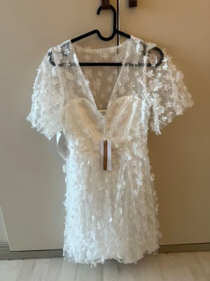 Vit studentklänning  - Superfin vit kortklänning med djup v-ringning och 3D-blommor över hela klänningen. Klänningen har korta ärmar i transparent tyg och markerad midja. Från bubbel room