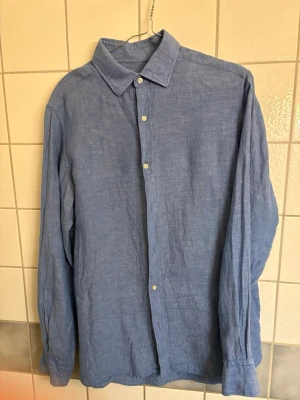 Ljus Blå linneskjorta med knappar - Snygg blå skjorta i linne från Massimo Dutti med klassisk krage och knäppning framtill. Skjortan har långa ärmar med en avslappnad passform. Perfekt för en stilren och enkel look.