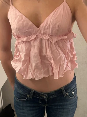 Ljusrosa volangig linnetopp - Supersöt ljusrosa linnetopp med tunna axelband och volangdetaljer framtill. Toppen har en luftig och flowy passform, perfekt för varma dagar. Materialet känns mjukt och lätt mot huden.