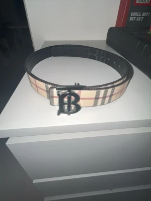 Burberry bälte med klassiskt rutmönster - Säljer ett Burberry bälte med det ikoniska beigea och bruna rutmönstret samt en stor metallspänne med Burberrys logotyp. Bältet kommer med originalpåse, box och kvitto. Perfekt accessoar för att lyfta din outfit. OBS DEFEKT PÅ SISTA BILD
