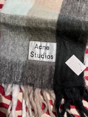 Rutig halsduk från Acne Studios - Mjuk och fluffig halsduk från Acne Studios i rutigt mönster med nyanser av svart, grått, beige och vitt. Stor logotyp framtill och långa fransar i ändarna. Perfekt accessoar för att lyfta din outfit.