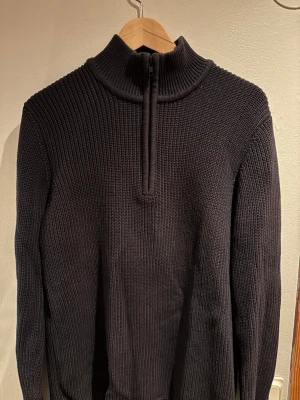 Svart stickad halvzip från Weekday - Mörk stickad tröja från Weekday med hög krage och halvzip-dragkedja. Grovstickad struktur och långa ärmar ger en avslappnad men stilren look. Perfekt för lager på lager och enkel att matcha med olika outfits.