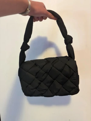 Svart handväska  - Trendig svart handväska med quiltat mönster och vadderad känsla. Väskan har en bred axelrem med snygga knutdetaljer på båda sidor. Jag har inte använt den och jag köpte den från Åhléns 💗