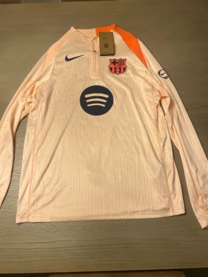 Barça långärmad träningströja Nike - Ljusrosa långärmad träningströja från Nike med FC Barcelona-logga och Spotify-sponsor på bröstet. Tröjan har en orange detalj vid kragen och tryck på ärmen. Materialet är lätt och andas, perfekt för fotbollsträning.