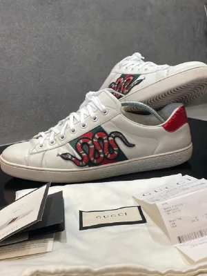 Gucci Ace sneakers  - Säljer ett par vita Gucci Ace sneakers i läder med ikonisk broderad orm på sidan och gröna/röda detaljer. Baksidan har metallicröd och metallicgrön hälflik i ormskinnslook. 