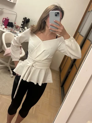 Vit omlottopp med bälte - Snygg vit omlottopp med bälte i midjan och volang nertill. Toppen har långa ärmar och kan bäras offshoulder för en trendig look. Perfekt att styla med jeans eller kjol.