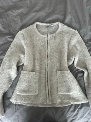 Beige fluffig kofta från Isolde - Mysig beige kofta från Isolde med fluffig textur och rund halsringning. Koftan har dragkedja framtill och två stora fickor. Perfekt för lager på lager och ger en avslappnad men ändå stilren look.