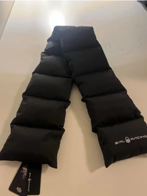 Svart quiltad halsduk Sail Racing - Snygg svart quiltad halsduk från Sail Racing. Halsduken är vadderad och har en modern, puffig design som håller dig varm under kyliga dagar. Perfekt accessoar för dig som gillar sportig och stilren look. Priset kan diskuteras 