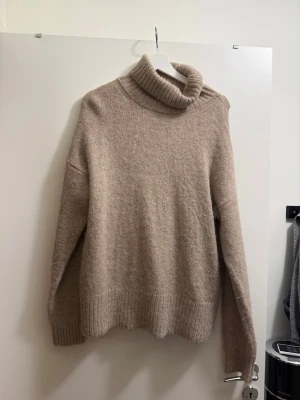 Beige polotröja i mjuk stickad kvalitet - Säljer en beige stickad polotröja med oversized passform och bred ribbad mudd vid ärmslut och nederkant. Tröjan har hög polokrage och är gjord i ett mjukt material som känns skönt mot huden. Perfekt för kalla dagar och enkel att styla. 39% ull 39% alpacka ull