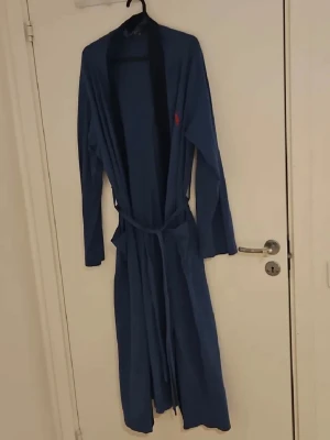 Blå morgonrock från Polo Ralph Lauren - Mysig blå morgonrock från Polo Ralph Lauren med klassisk omlottmodell och knytskärp i midjan. Lång ärm och diskret röd logga broderad på bröstet. Perfekt för chilliga morgnar eller kvällar hemma. Tillverkad i mjukt tyg för extra komfort. Knappt använd!.