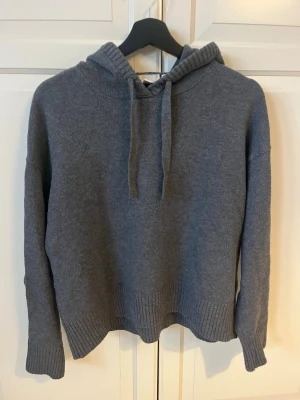 Mörkgrå stickad hoodie - Mysig mörkgrå stickad hoodie med huva och dragsko. Tröjan har ribbade muddar vid ärmslut och nederkant samt en avslappnad passform. Perfekt för en chill och avslappnad stil. Materialet känns mjukt och skönt mot huden.