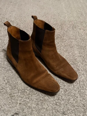 Bruna mocka Chelsea boots - Snygga Chelsea boots i brun mocka med klassiska svarta resårpaneler på sidorna och låg klack. Bootsens spetsiga tå ger en stilren look och de har dragflikar bak för enkel påtagning. Perfekta för dig som vill ha en clean och trendig stil.
