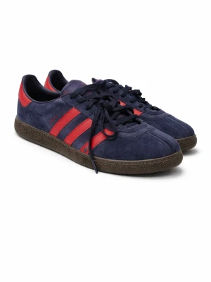 Adidas München blå och röda sneakers - Snygga Adidas Gazelle sneakers i mörkblå mocka med klassiska röda ränder och detaljer. Skorna har svart snörning och en brun platt sula med retrovibbar. Perfekta för dig som vill ha en ikonisk och stilren sneaker med streetkänsla.dessa är svåra att få tag på