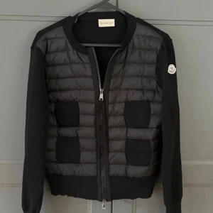 Svart kofta från Moncler - Säljer en svart quiltad kofta från Moncler med ribbade muddar och fyra fickor framtill. Tröjan har långärmade stickade ärmar, vadderad framsida och dragkedja hela vägen. Klassisk Moncler-logga på ärmen och snygg, stilren look.