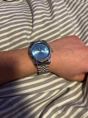 Rolle klocka  - Bilderna visar en silverfärgad klocka med blå urtavla och metallänk, men ingen väst syns.