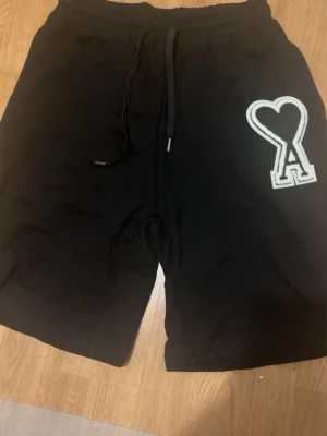 Svarta shorts från Ami Paris - Svarta shorts från Ami Paris med snörning i midjan och stor vit hjärtformad A-logga på ena benet. Sköna och stilrena, perfekta för sommaren. Tillverkade i mjuk bomull för maximal komfort.