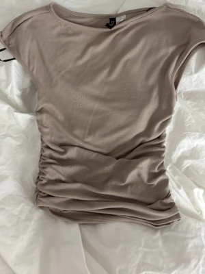 Beige topp med rynkade sidor - Snygg beige topp från H&M med rynkade sidor som ger en cool och smickrande passform. Toppen har korta ärmar och är tillverkad i mjukt bomullsmaterial. Perfekt att styla med jeans eller kjol för en trendig look. Lite nopprig på vissa ställen se bilderna