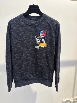 Mörkgrå sweatshirt från Dsquared2 - Cool mörkgrå sweatshirt från Dsquared2 med broderade patches på bröstet, bland annat en amerikansk flagga och texten 'ICON CATEN'. Tröjan har rund halsringning och långa ärmar, perfekt för en avslappnad streetstyle.
