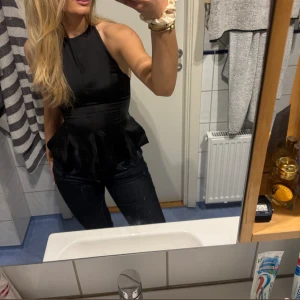 Svart halterneck topp med volang - Snygg svart halterneck topp med volangdetaljer i midjan. Toppen har en tight passform upptill och är ärmlös, vilket ger en stilren och trendig look. Perfekt att matcha med jeans eller kjol för en cool outfit.