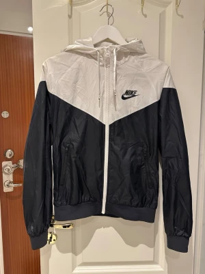 Svartvit vindjacka från Nike - Nike vindjacka i svart och vitt med ikonisk logga på bröstet. Jackan har dragkedja framtill, två fickor med dragkedja och en huva. Tillverkad i lätt, vindtätt material och har muddar vid ärmslut och nederkant. Perfekt för en sportig och avslappnad stil. En del slitningar, se bilder 