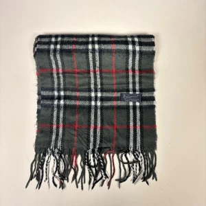 Rutig halsduk från Burberrys i ull - Klassisk rutig halsduk från Burberrys i 100% lammull. Mörkgrå bas med röda, vita och svarta ränder samt fransar i ändarna. Perfekt accessoar för att lyfta din outfit med en tidlös brittisk stil.