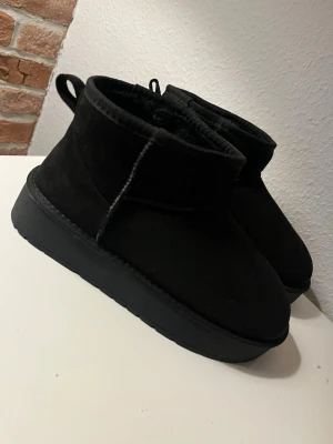 Svarta fluffiga tofflor med platåsula - Säljer ett par svarta tofflor med hög platåsula och mjukt, fluffigt foder. Tofflorna har en enkel design med rund tå och dragflik bak. Perfekta för att hålla fötterna varma och sköna hemma. Materialet är mockaliknande på utsidan och mjukt foder inuti.