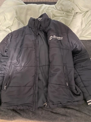 Svart pufferjacka från Lotto - Svart pufferjacka från Lotto med quiltad design och dragkedja framtill. Jackan har två fickor med dragkedja och hög krage. Perfekt för kyliga dagar och enkel att matcha med olika outfits.