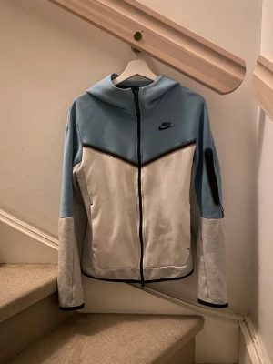Nike Tech Fleece hoodie - Nike hoodiejacka i storlek XS i blå, vitt och grått. Jackan har hel dragkedja, huva och svart Nike-logga på bröstet. Snygg kontrast med svarta detaljer och ficka med dragkedja på ärmen. 