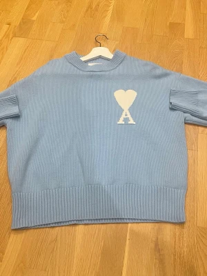 Ljusblå stickad tröja från Ami Paris - Säljer en ljusblå stickad tröja från Ami Paris med deras ikoniska vita hjärta och A på bröstet. Tröjan har sköna ribbade muddar och en avslappnad passform. Perfekt för dig som gillar stilrena och trendiga plagg. Är öppen för prisförslag vid snabb och smidig affär 🙌