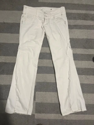 Vita bootcut jeans med detaljer - Säljer ett par vita bootcut jeans med fransade detaljer längs benen och dubbla knappar i midjan. Jeansen har fem fickor och dekorativa nitar. Perfekta för dig som gillar en unik och trendig look.