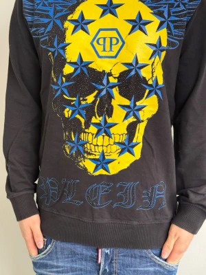 Philipp Plein hoodie med döskalle - Svart hoodie från Philipp Plein med stor gul döskalle och blå stjärnor på framsidan. Loggan och texten 'PLEIN' syns tydligt. Hoodien har huva och ribbade muddar. Materialet känns mjukt och bekvämt, perfekt för dig som gillar streetwear och statement-plagg. Han på bild är 175cm och väger 74kg