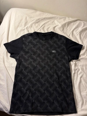 Svart mönstrad t-shirt från BOSS - Snygg svart t-shirt från BOSS med diskret rutmönster framtill och liten logga på bröstet. Klassisk rund halsringning och korta ärmar. Perfekt för en stilren och modern look.