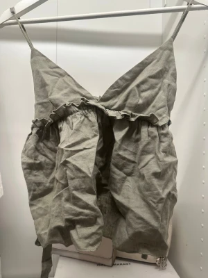 Grön linnetopp med volang och knyt - Snygg ljusgrön linnetopp med smala axelband, v-ringad hals och volangdetalj under bysten. Toppen har knyt i sidorna för justerbar passform och är croppad. Perfekt för varma dagar och enkel att styla med jeans eller kjol.