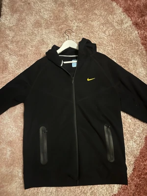 Svart Nike hoodie med gul logga - Snygg svart hoodie från Nike med dragkedja framtill och två fickor med dragkedja. På bröstet sitter en gul Nike Swoosh. Hoodien har huva och är tillverkad i ett mjukt material som känns bekvämt. Perfekt för en sportig och avslappnad stil.