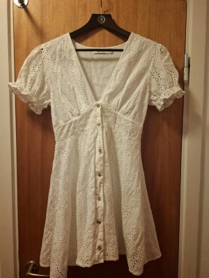 Vit kort klänning NA-KD - Söt vit kort klänning från NA-KD med broderade spetsdetaljer och knappar framtill. Klänningen har v-ringning, puffiga korta ärmar. Perfekt som studentklänning. 