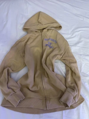 Beige Powerpuff Girls hoodie - Beige hoodie med dragkedja och huva, dekorerad med Powerpuff Girls-tryck på ryggen i färgglada nyanser och texten 'GIRLS UNITE'. På bröstet finns ett mindre tryck i lila. Mjukt material och breda muddar vid ärmslut och nederkant.