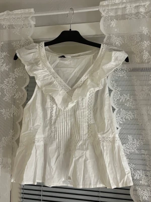 Vit volangblus med spetsdetaljer - Söt vit ärmlös blus med volang runt V-ringningen och fina spetsdetaljer längs framstycket. Blusen har veckade detaljer framtill och en lätt utsvängd passform. Perfekt för en romantisk och somrig look. Tillverkad i ett tunt, luftigt tyg.