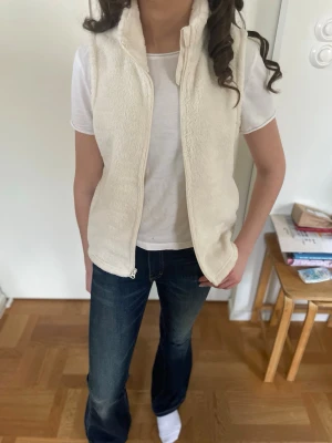 Krämvit fleeceväst med dragkedja - Säljer en krämvit fleeceväst med hög krage och dragkedja framtill. Västen är gjord i mjukt, fluffigt material som ger en mysig känsla och har en rak passform. Perfekt för dig som vill ha en enkel men snygg look. Fin att bära inomhus t.ex. till skolan eller som ett extra lager för värme. Står storlek xl men tror att det är barnstorlek för sitter som xs kanske s. 