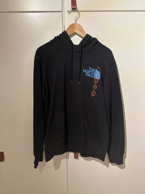Svart hoodie från The North Face - Svart hoodie från The North Face med stor blå logga och orange grafiskt tryck både fram och bak. Klassisk känguruficka och justerbar huva med snören. Tillverkad i mjukt bomullsmaterial, perfekt för chill dagar eller när du vill ha en clean streetwear-look.