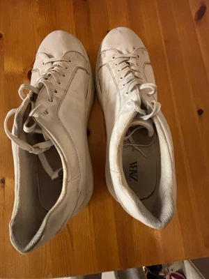 Vita sneakers från Zara i skinn hyfsat bra skick  - Stilrena vita sneakers från Zara i storlek 46. Skorna har klassisk design med rund tå, snörning och platt sula. Tillverkade i skinn för en clean look som passar till det mesta. Perfekta för dig som gillar minimalistisk stil.