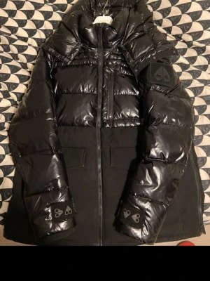 Svart pufferjacka Moose Knuckles - Säljer en svart pufferjacka från Moose Knuckles med glansig finish och coola logodetaljer på ärmar och bröst. Jackan har hög krage, dragkedja framtill och stora fickor. Perfekt för kalla dagar och riktigt snygg streetstil.