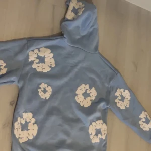 Denim tears Hoodie  - Snygg ljusblå hoodie med vita blommönster över hela tröjan. Hoodien har en klassisk huva och är tillverkad i mjukt material, perfekt för en chill och avslappnad stil. Passar dig som gillar streetwear och vill sticka ut lite extra.