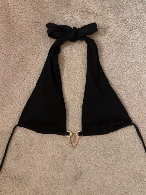 Svart bikiniöverdel med guldkedjor - Snygg svart bikiniöverdel i halterneck-modell med dekorativa guldfärgade kedjor och gnistrande stenar framtill. Bikinin har breda band som knyts i nacken och smala knytband i ryggen. Materialet är ribbat för extra stilpoäng.